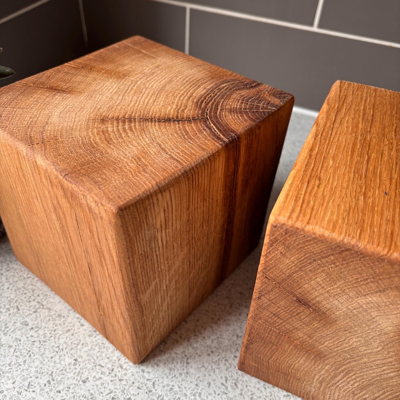 May include: Two solid wooden blocks with a natural light brown finish, showcasing a prominent wood grain pattern and visible growth rings on the top surfaces. The blocks are cubic in shape with smooth, straight edges and a warm, earthy tone.