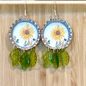 May include: A pair of sunflower earrings with green glass leaves dangling from the bottom. The earrings are made from bottle caps with a white background and a sunflower image.