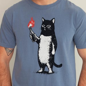 May include: Blue jean coloured t-shirt featuring a black and white cat holding a bottle with a red flame. The cat has a serious expression. The shirt is a solid colour and has short sleeves.