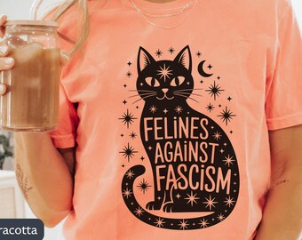 May include: Terracotta-colored t-shirt featuring a black cat design with the words "Felines Against Fascism." The cat is surrounded by stars and a crescent moon. The shirt is a solid color and has short sleeves.