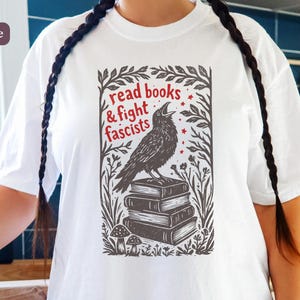 以下が含まれることがあります： 本の上にカラスが描かれた白いTシャツ。赤い文字で「read books & fight fascists」と書かれています。ゆったりとしたフィット感のカジュアルなTシャツです。
