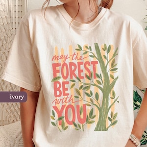 May include: Ivory t-shirt featuring the phrase "may the FOREST BE with YOU" in coral and green, with a tree illustration. The design includes yellow vertical stripes. The shirt is a relaxed, casual style.
