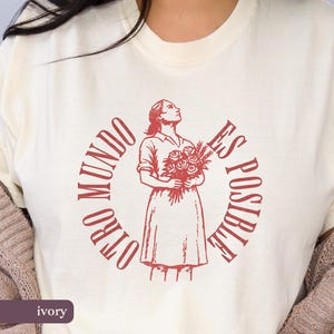Peut inclure: T-shirt ivoire avec une illustration rouge d'une femme tenant des fleurs et le texte "OTRO MUNDO ES POSIBLE" dans un cercle. Le t-shirt a un col rond et des manches courtes.