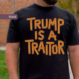 May include: Black crew neck t-shirt featuring the orange text "TRUMP IS A TRAITOR". The shirt is made of a soft material. The word "black" is in a purple box in the upper left corner. The shirt is a casual style.