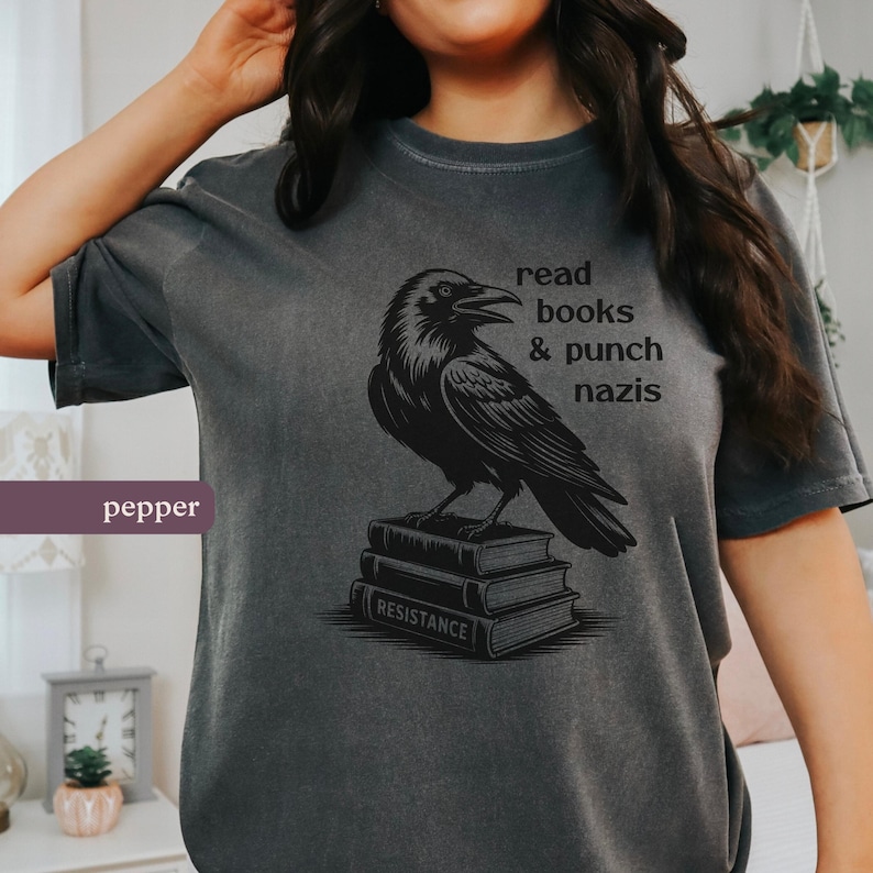 Op de afbeelding: Donkergrijs t-shirt met een zwarte ravenillustratie op een stapel boeken. De tekst "read books & punch nazis" is op de shirt gedrukt. Het woord "resistance" staat op het onderste boek.