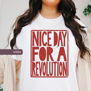 May include: White t-shirt featuring a red rectangular graphic with the text "NICE DAY FOR A REVOLUTION". The text is in a bold, distressed font. The shirt is a crew neck style.