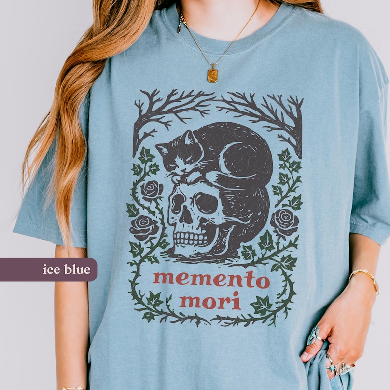 May include: Pale blue t-shirt with a gothic design. The graphic features a skull with a cat resting on top, framed by roses and vines. The words "memento mori" are printed below. The shirt has short sleeves.