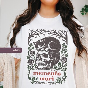 May include: White t-shirt with a black cat resting on a skull, the words "memento mori" in red. The design includes tree branches, roses, and green vines. A casual wear item.