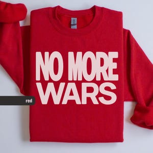 May include: A red sweatshirt featuring the text "NO MORE WARS" in large, white capital letters. The garment is folded, with the sleeves visible. The word "red" is in a black rectangle in the lower left corner.