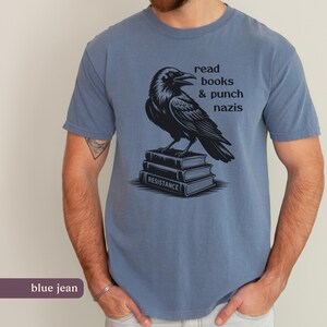 Op de afbeelding: Blauw spijkerstof T-shirt met een zwarte raven illustratie op een stapel boeken. De boeken zijn voorzien van het woord "RESISTANCE". Het shirt heeft ook de tekst "read books & punch nazis" erop.