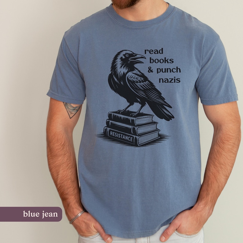 Op de afbeelding: Blauw spijkerstof T-shirt met een zwarte raven illustratie op een stapel boeken. De boeken zijn voorzien van het woord "RESISTANCE". Het shirt heeft ook de tekst "read books & punch nazis" erop.