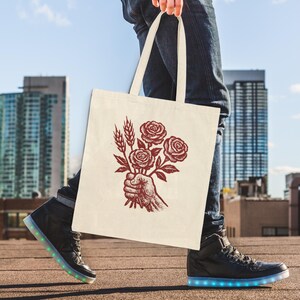 May include: A cream tote bag with a red illustration of a hand holding roses and wheat. The bag is carried by a person wearing blue jeans and black high-top trainers with illuminated soles. The background shows city buildings and a blue sky.