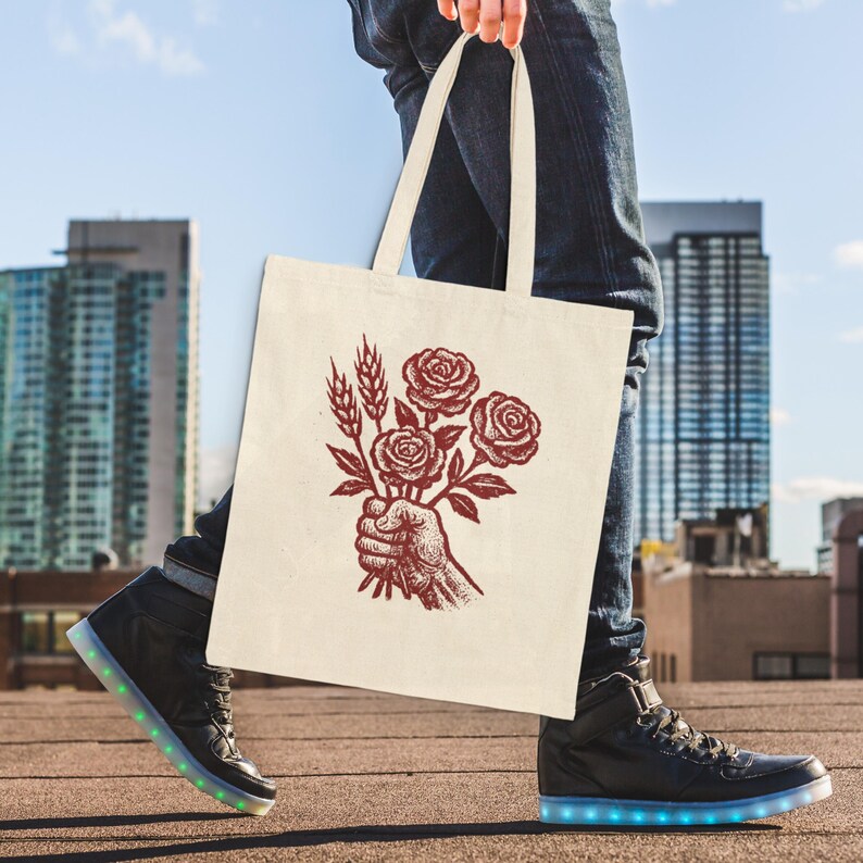 May include: A cream tote bag with a red illustration of a hand holding roses and wheat. The bag is carried by a person wearing blue jeans and black high-top trainers with illuminated soles. The background shows city buildings and a blue sky.