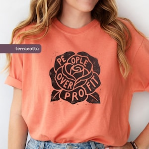 May include: Terracotta-colored t-shirt with a black rose graphic. The rose design features the words "PEOPLE OVER PROFIT" in a circular arrangement. The shirt has short sleeves and a relaxed fit.