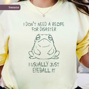 Può includere: T-shirt giallo chiaro con la scritta "I DON'T NEED A RECIPE FOR DISASTER" e l'illustrazione di una rana. Sotto, la frase "I USUALLY JUST EYEBALL IT". La maglietta ha una vestibilità ampia.