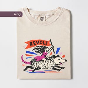 May include: An ivory t-shirt featuring a graphic print of a person riding a large opossum while holding a red flag with the text REVOLT. The illustration uses black, pink, blue, and red colours, and the shirt has a Comfort Colors brand neck label.