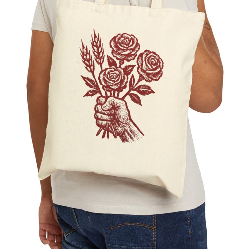 May include: A cream-coloured tote bag with a burgundy illustration of a hand holding roses and wheat. The design is a vintage-style drawing, suitable for carrying groceries or books. The bag is made of a durable fabric.