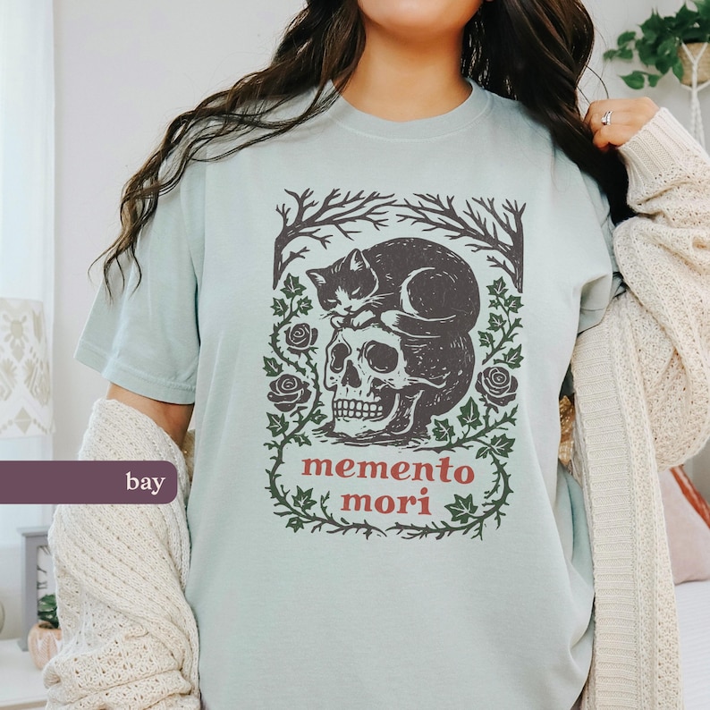 May include: A pale blue t-shirt with a skull and a sleeping cat design, framed by roses and vines. The words "memento mori" are printed in red. The shirt has short sleeves.