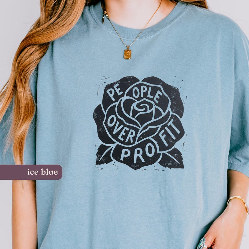 May include: Ice blue t-shirt with a black rose graphic. The rose design features the words "People Over Profit" in a circular arrangement. The shirt is a solid color and has short sleeves.