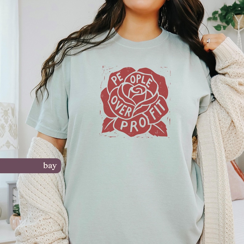 May include: A light blue t-shirt with a red rose graphic. The rose design features the words "People Over Profit" in a circular arrangement. The shirt is a solid color and has short sleeves.