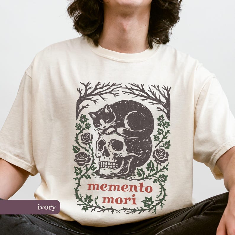 May include: Ivory t-shirt with a graphic print of a black cat on a skull, with the words "memento mori" in red. The design includes dark green floral and tree branch elements. The shirt is a neutral colour.