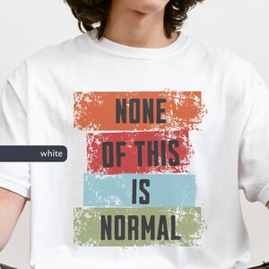 May include: White t-shirt featuring the slogan "NONE OF THIS IS NORMAL" in black, block lettering. The text is set against four horizontal, distressed-effect rectangles in orange, red, blue, and green. A casual style t-shirt.