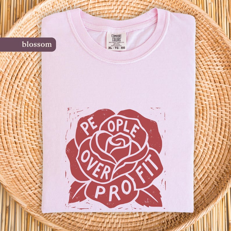 May include: A light pink Comfort Colors t-shirt with a crew neck. The shirt features a red rose graphic with the words "PEOPLE OVER PROFIT" in white. The shirt is folded on a woven basket.