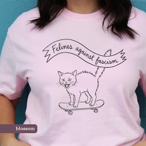 Könnte beinhalten: Hellrosa T-Shirt mit einer schwarzen Grafik einer Katze auf einem Skateboard, mit dem Text "Felines against fascism" in einem Banner darüber. Das Wort "blossom" steht unten auf dem Shirt.