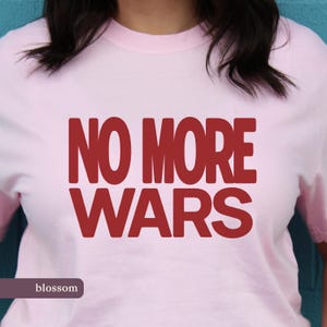 May include: A pale pink t-shirt featuring the text "NO MORE WARS" in large, red letters. The word "blossom" is printed in a smaller font at the bottom of the shirt.
