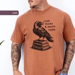 Op de afbeelding: Een roestkleurig t-shirt met een zwarte raaf bovenop een stapel boeken. De tekst luidt "read books & punch nazis". Het woord "RESISTANCE" staat op het onderste boek.