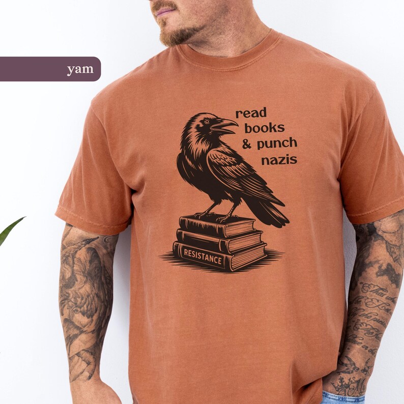 Op de afbeelding: Een roestkleurig t-shirt met een zwarte raaf bovenop een stapel boeken. De tekst luidt "read books & punch nazis". Het woord "RESISTANCE" staat op het onderste boek.