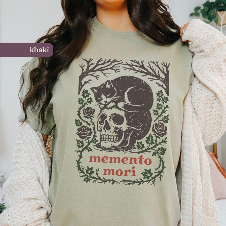May include: Khaki t-shirt with a graphic design of a skull topped by a cat, framed by roses and vines. The words "memento mori" are printed in red. A casual wear item.