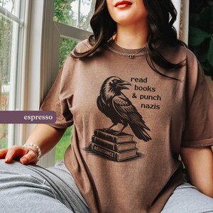 Op de afbeelding: Espresso-kleurig T-shirt met een zwarte ravenillustratie bovenop boeken. Tekst boven de raaf luidt "read books & punch nazis". Het onderste boek toont "RESISTANCE".