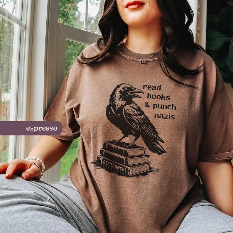 Op de afbeelding: Espresso-kleurig T-shirt met een zwarte ravenillustratie bovenop boeken. Tekst boven de raaf luidt "read books & punch nazis". Het onderste boek toont "RESISTANCE".