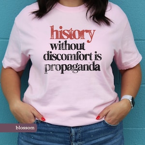 Pode incluir: T-shirt rosa claro com a frase "history without discomfort is propaganda" em vermelho e preto. A t-shirt é feita de um material macio e é um artigo de roupa casual.