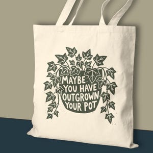 May include: Cream-colored tote bag with a dark green illustration of a plant in a pot, surrounded by leaves. The text "MAYBE YOU HAVE OUTGROWN YOUR POT" is printed on the bag. The bag has two long handles.