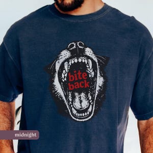 Può includere: T-shirt blu notte con grafica di una testa di lupo ringhiante e la scritta rossa "bite back". La bocca del lupo è aperta, mostrando i denti. La maglietta è a tinta unita e ha un girocollo.