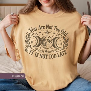 May include: A mustard yellow short-sleeve t-shirt featuring a dark brown graphic print of a moon cycle with stars and leaves. The text reads, "You Are Not Too Old & IT IS NOT TOO LATE" in a decorative font surrounding the central moon illustration.