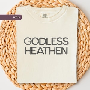 May include: Ivory-colored t-shirt with the words "GODLESS HEATHEN" printed in dark gray block letters. The shirt is made of a soft material and is displayed on a woven, light brown surface.