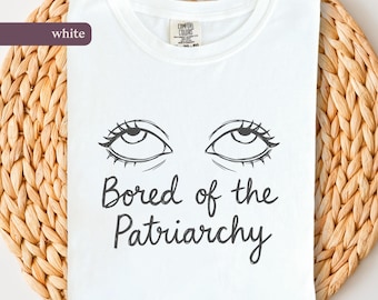 May include: White t-shirt with the text "Bored of the Patriarchy" in a handwritten font. The shirt features a graphic of two eyes looking upwards with long eyelashes. The shirt is on a woven, beige surface.