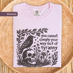 Pode incluir: Camiseta rosa claro com um gráfico preto de um corvo em um crânio, cercado por folhagens e cogumelos. O texto diz "you cannot comply your way out of tyranny".