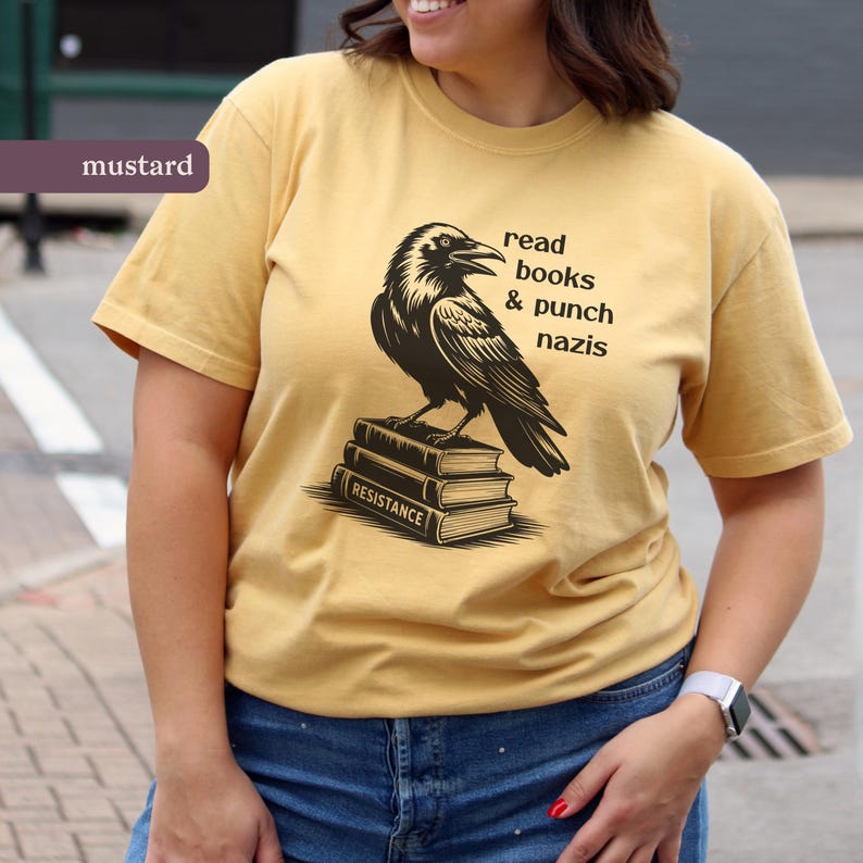 Op de afbeelding: Mosterdkleurig T-shirt met een zwarte raaf op een stapel boeken. De boeken zijn gemarkeerd met "RESISTANCE". De tekst op het shirt luidt "read books & punch nazis."