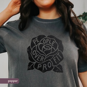 May include: Dark gray t-shirt with a black rose graphic. The rose design features the words "PEOPLE OVER PROFIT" in a circular arrangement. The shirt is a solid color and has a vintage, worn look.