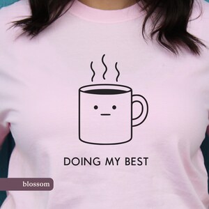 Op de afbeelding: Lichtroze t-shirt met een zwarte afbeelding van een koffiemok met stoom. De tekst "DOING MY BEST" staat onder de mok. Het woord "blossom" staat in een donkerpaarse rechthoek onderaan.