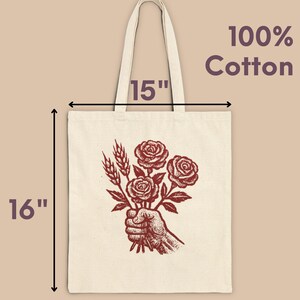 May include: Cream tote bag featuring a red illustration of a hand holding roses and wheat. The bag measures 40.6 cm tall and 38.1 cm wide. The text "100% Cotton" is printed on the upper right.