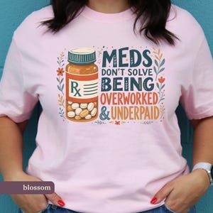 May include: A light pink t-shirt featuring a graphic of an orange prescription bottle filled with white pills. The text reads: MEDS DON'T SOLVE BEING OVERWORKED & UNDERPAID. The design is framed by colourful floral vines in shades of blue, orange, and green.