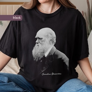 Könnte beinhalten: Schwarzes T-Shirt mit einem Graustufen-Porträt von Charles Darwin, erkennbar an seinem langen weißen Bart. Der Name "Charles Darwin" ist darunter in Schreibschrift geschrieben. Das T-Shirt ist einfarbig schwarz.
