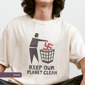 Puede incluir: Camiseta de color marfil con un gráfico de una persona tirando una esvástica roja a una papelera. El texto "KEEP OUR PLANET CLEAN" está debajo del gráfico. La camiseta está hecha de un material suave.