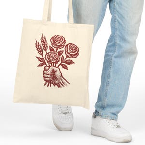 May include: Cream tote bag with a burgundy illustration of a hand holding roses and wheat. The bag is made of a sturdy fabric and has two handles. The design is a vintage-style graphic, suitable for shopping or carrying items.