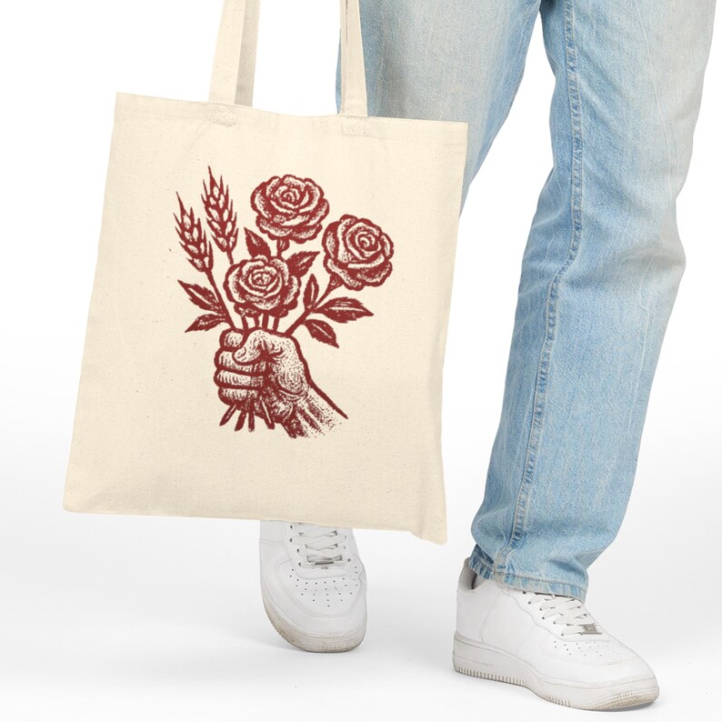 May include: Cream tote bag with a burgundy illustration of a hand holding roses and wheat. The bag is made of a sturdy fabric and has two handles. The design is a vintage-style graphic, suitable for shopping or carrying items.